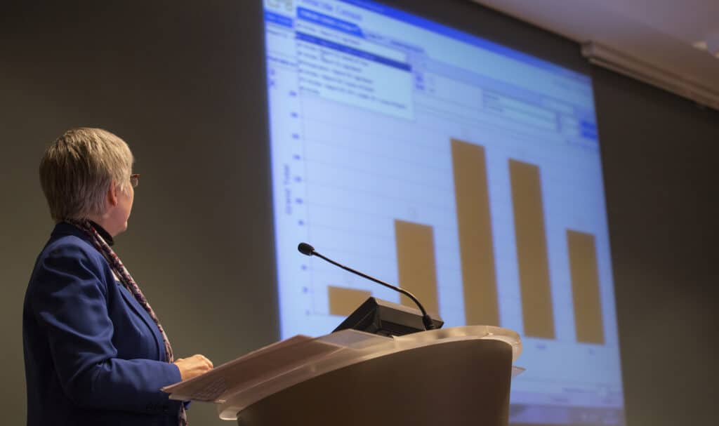 Woman standing at a microphone looking up to a projector