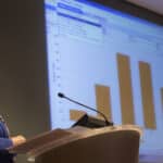Woman standing at a microphone looking up to a projector