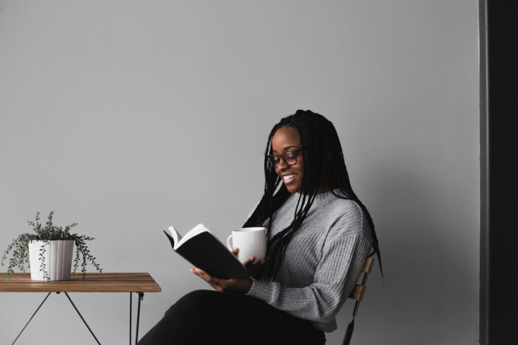 A woman smiling and sitting at a table holding a white mug and reading a book