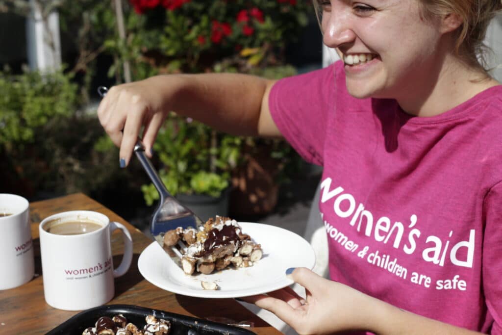 Woman wearing a pink Women's Aid t-shirt holding a plate with a slice of baked goods with Women's Aid mugs on the table