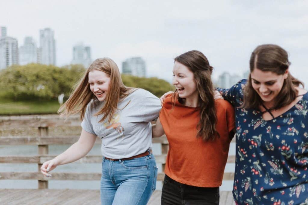 Three women with their arms over one another smiling with a a cityscape in the background