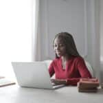 Woman smiling while sitting at a desk surrounded by books and looking at a laptop