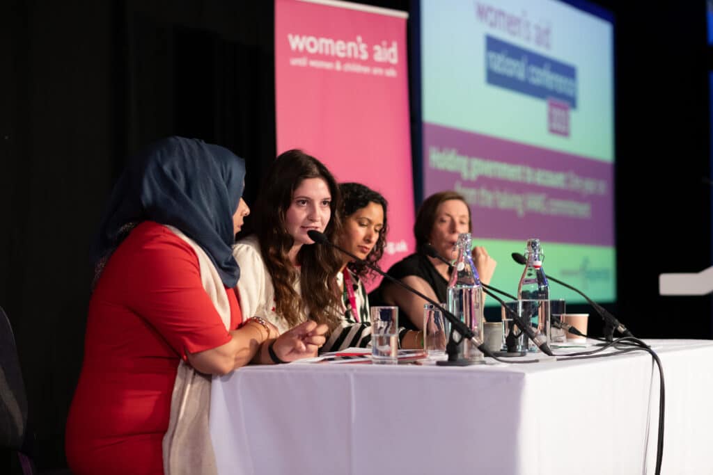 A panel of women with a Women's Aid banner and screen behind them