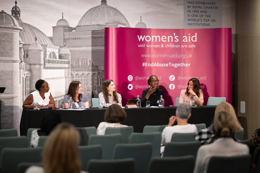 An audience and a panel of women sitting in front of a Women's Aid banner