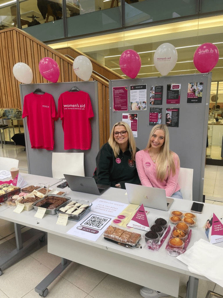 Two women smiling sitting at a table with baked goods and behind them boards with Women's Aid merch